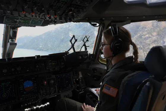 A young Air Force officer at the controls of a C-17 cockpit above mountainous terrain.