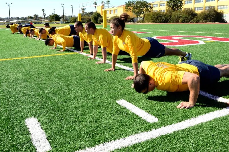Navy ROTC students performing pushups in a line during a fitness assessment on a turf field