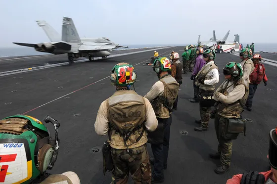 An F/A-18 Hornet launching from a carrier flight deck as Navy crew members look on.