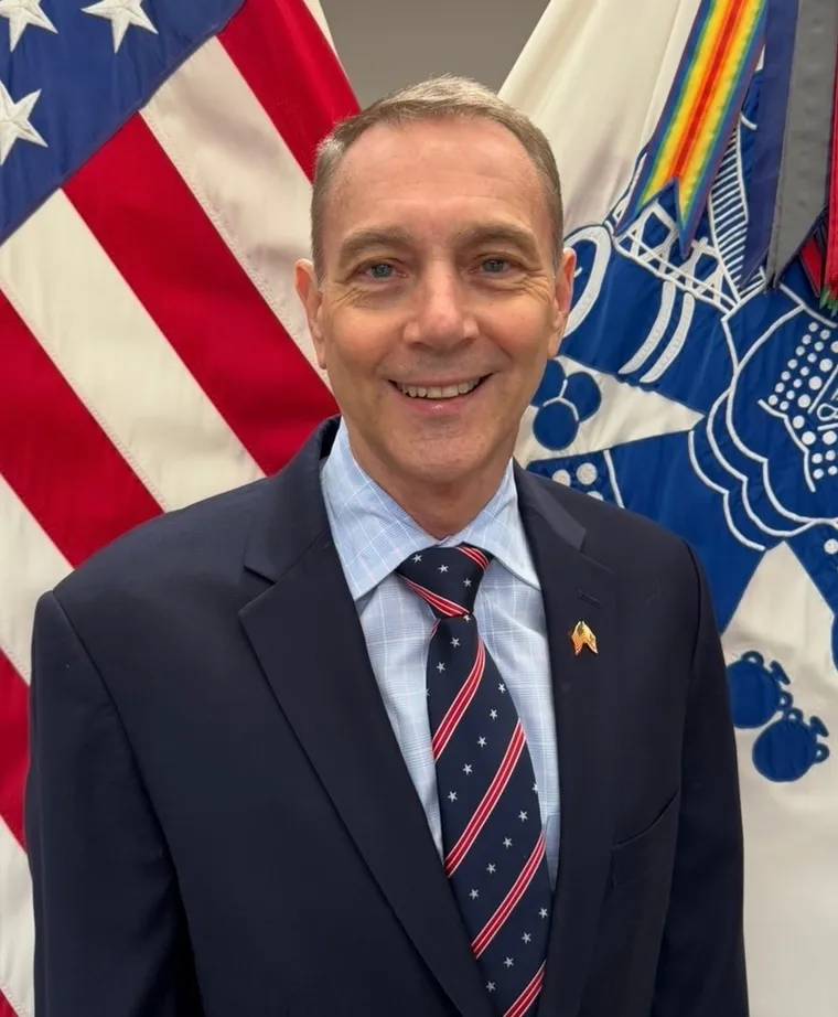 COL Lee Reynolds, U.S. Army, Retired, in a dark suit in front of the United States flag and the Army flag